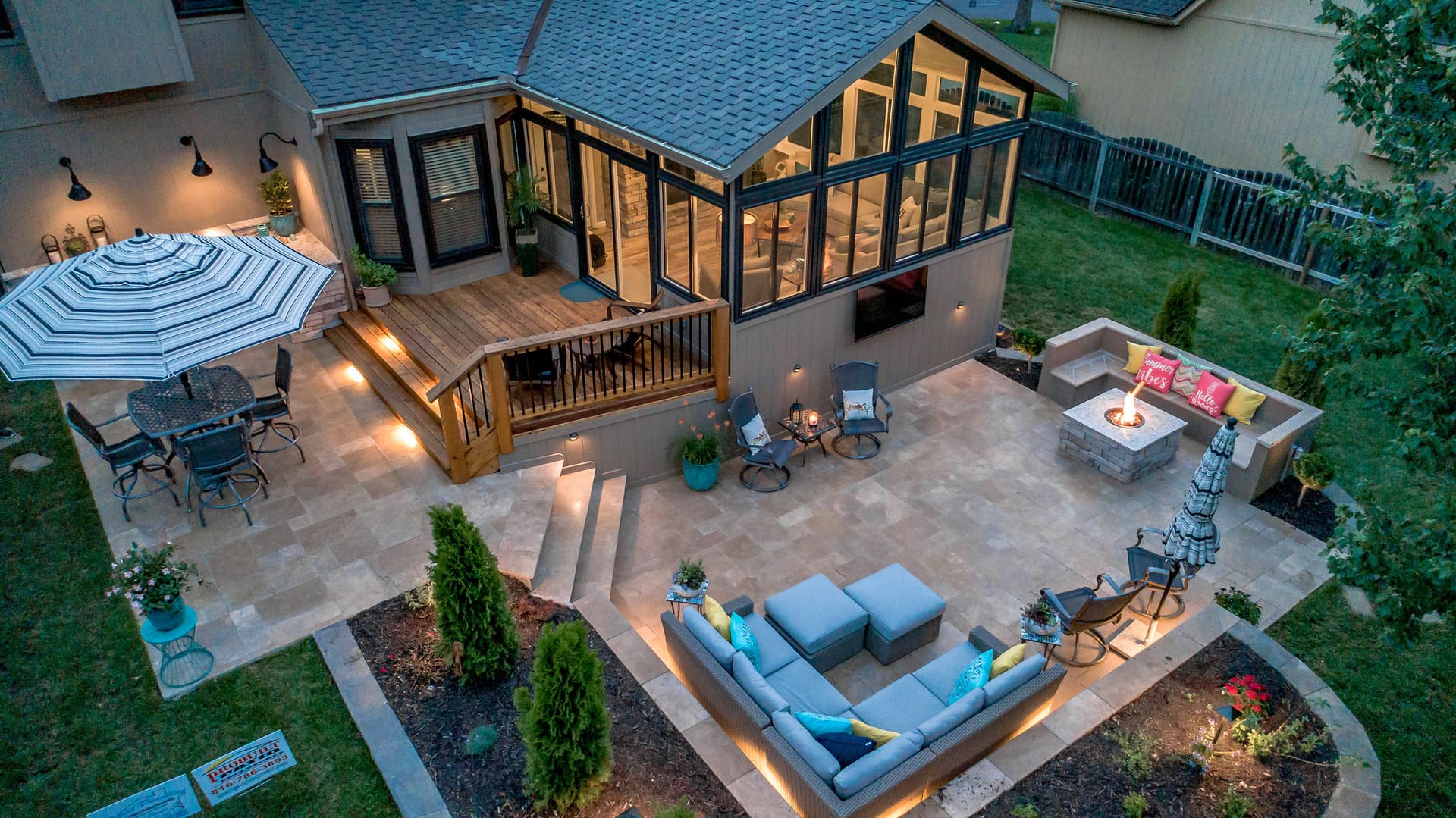 Aerial dusk view of a sunroom addition, wood deck, and paver patio with a fire pit and sunken lounge seating